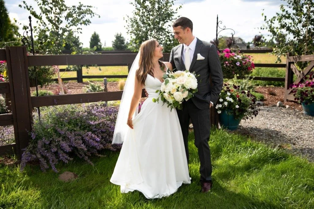 Bride and groom share a joyful glance while standing on lush green grass surrounded by lavender blooms and white roses at Mountain View Manor wedding venue in Enumclaw Washington near Seattle