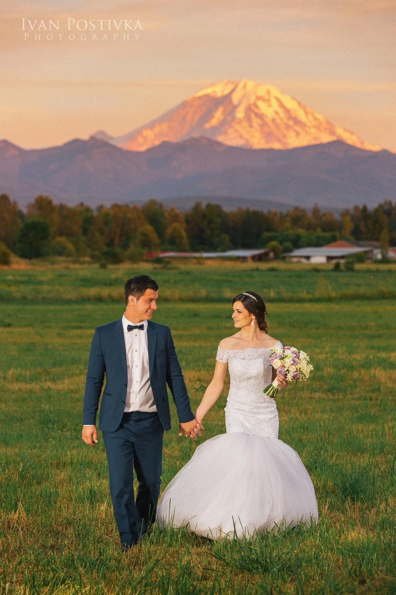 Bride and groom walking hand in hand through a green field at golden hour with Mount Rainier glowing at sunset behind them at Mountain View Manor wedding venue in Enumclaw Washington, photographed by Ivan Postivka Photography