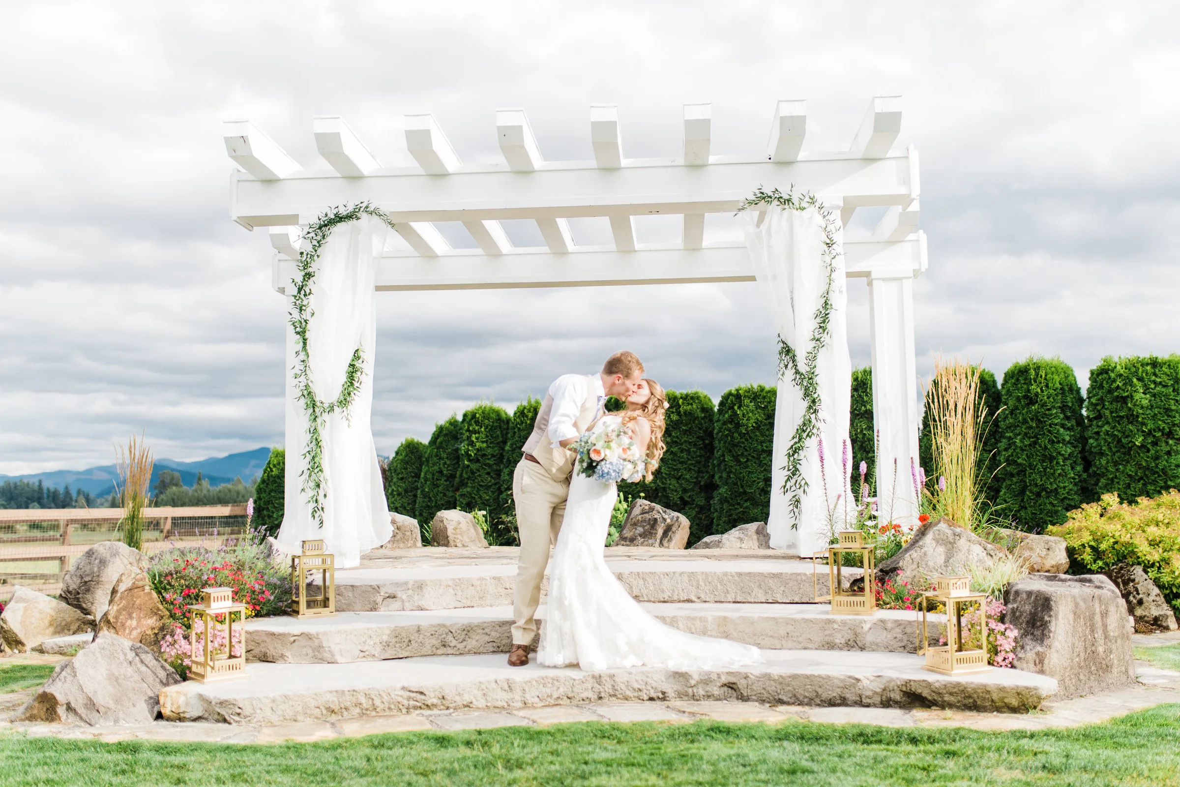 outdoor ceremony pergola kiss mountain view manor enumclaw - Mountain View Manor Bride and groom sharing a first kiss under a white pergola draped with greenery and white fabric at Mountain View Manor wedding venue in Enumclaw Washington with mountain views in the background