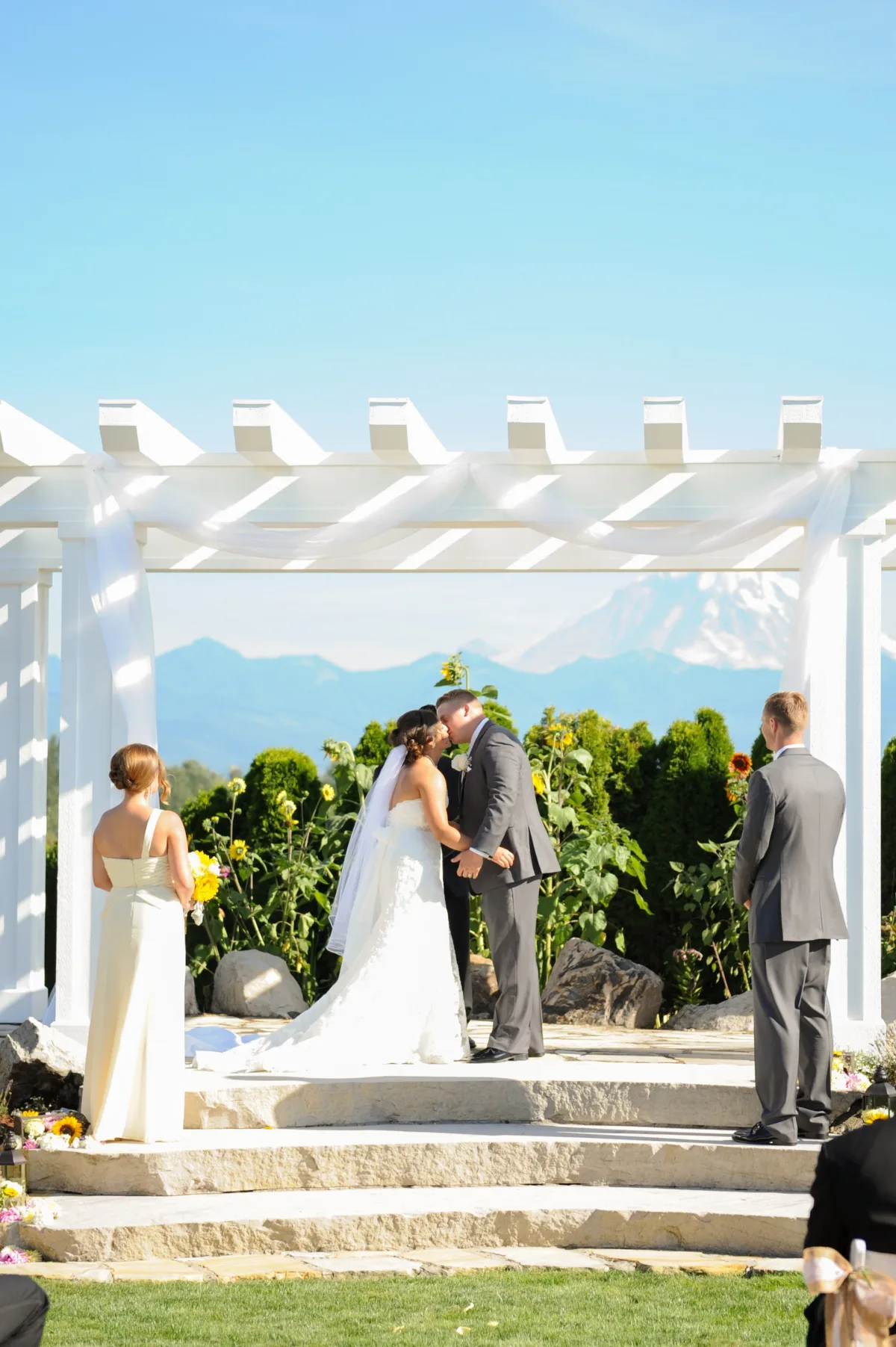 Bride and groom share their first kiss under a white pergola with Mount Rainier in the background surrounded by sunflowers at Mountain View Manor wedding venue in Enumclaw Washington
