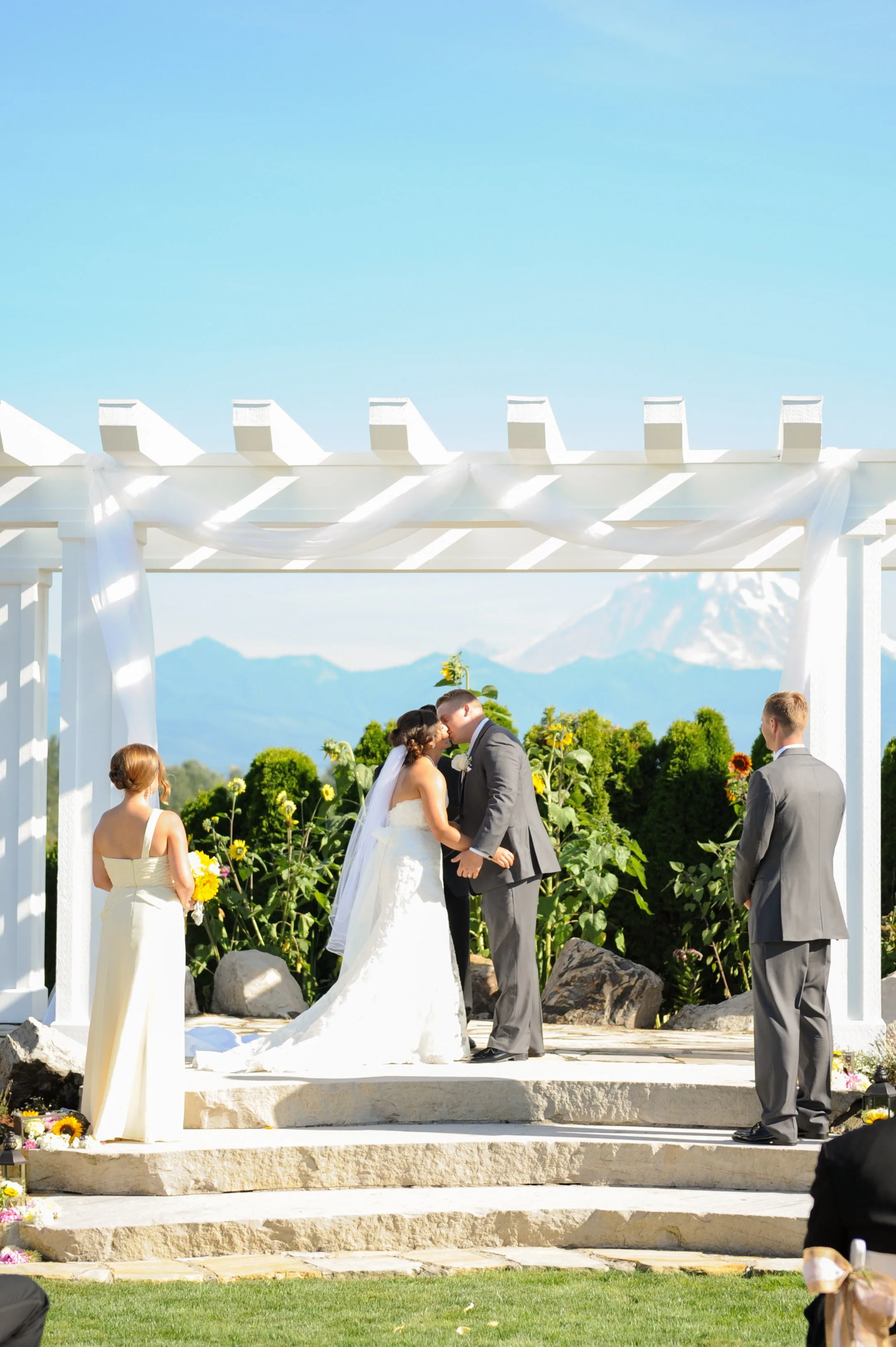 outdoor wedding ceremony first kiss mount rainier view - Packages & Pricing Bride and groom share their first kiss under a white pergola with Mount Rainier in the background surrounded by sunflowers at Mountain View Manor wedding venue in Enumclaw Washington