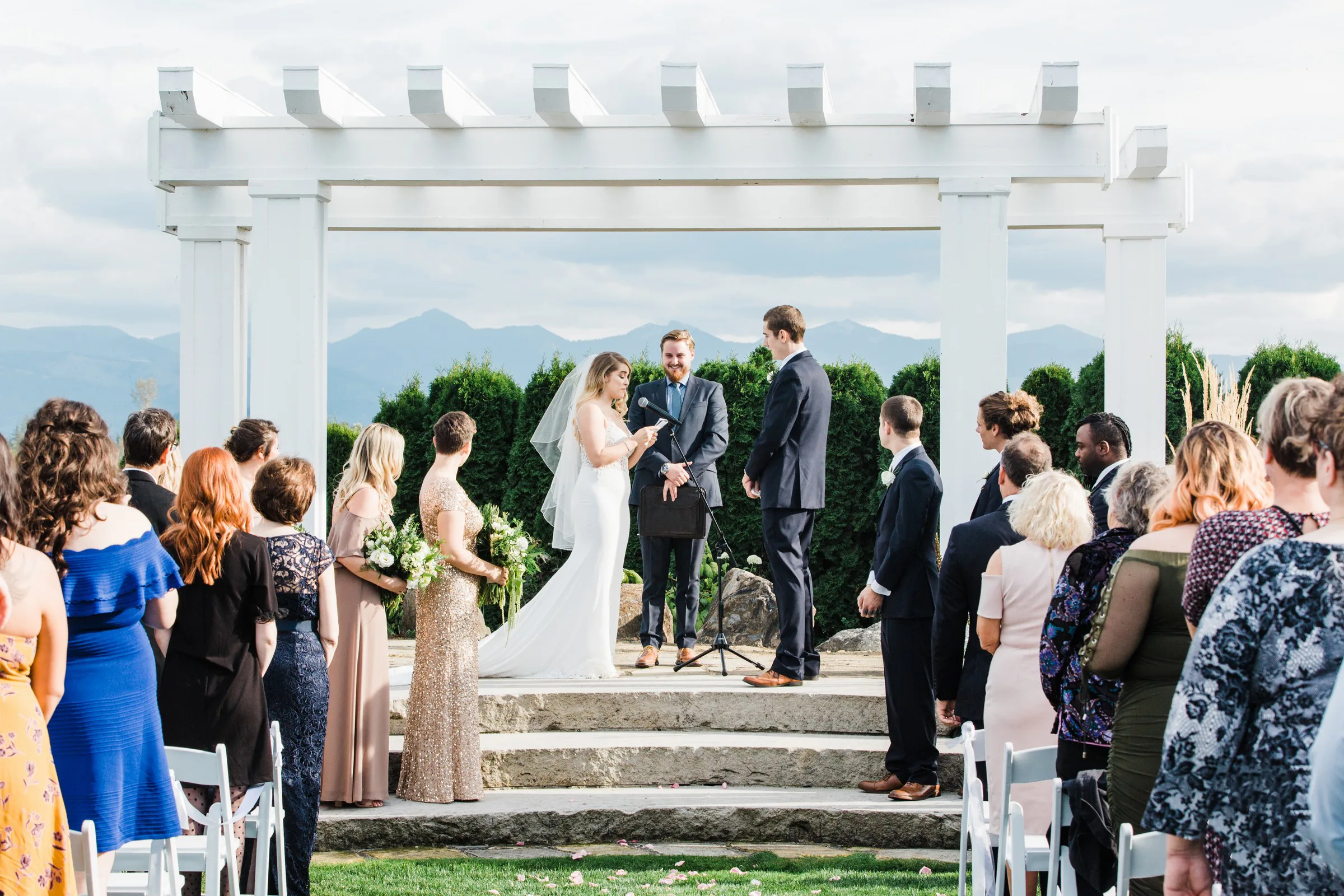 outdoor wedding ceremony mountain view manor enumclaw washington - Mountain View Manor Bride reading vows to groom under a white pergola with sweeping mountain views at Mountain View Manor wedding venue in Enumclaw Washington near Seattle