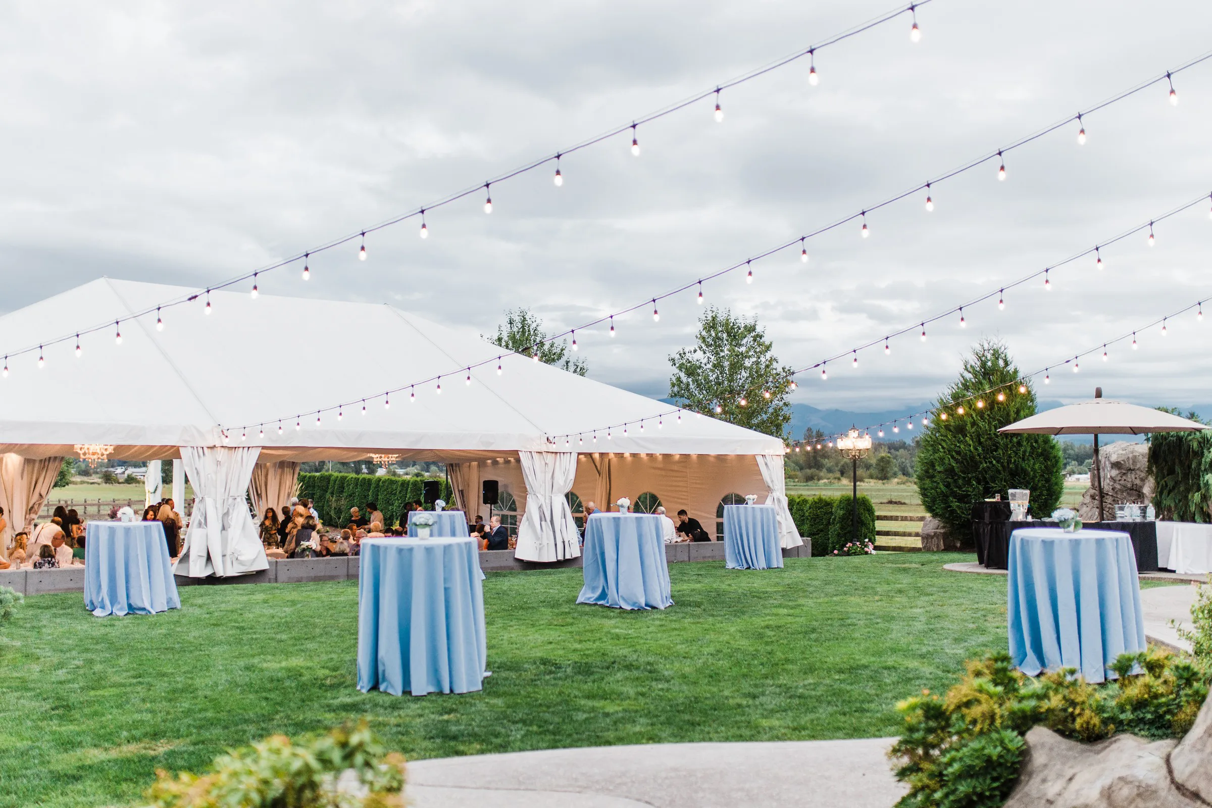 outdoor wedding reception tent string lights mountain view manor - Private Events Elegant outdoor wedding reception at Mountain View Manor in Enumclaw Washington featuring white frame tents, soft blue cocktail tables on green lawn, and bistro string lights strung overhead against a cloudy mountain backdrop